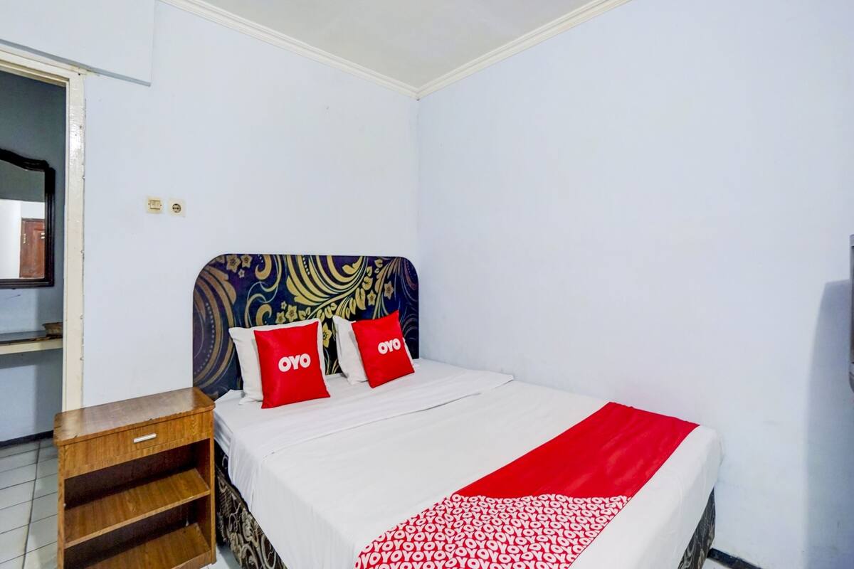 Een net opgemaakt bed met een rode deken en kussens, een houten nachtkastje en een hoofdbord met een patroon.