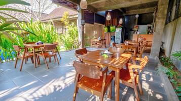 Un área de comedor al aire libre con mesas y sillas de madera, rodeada de exuberante vegetación y un suelo de baldosas.