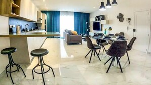 Une cuisine moderne avec un îlot central, un coin repas avec une table ronde et des chaises, un salon avec un canapé et des rideaux, et un téléviseur fixé au mur.