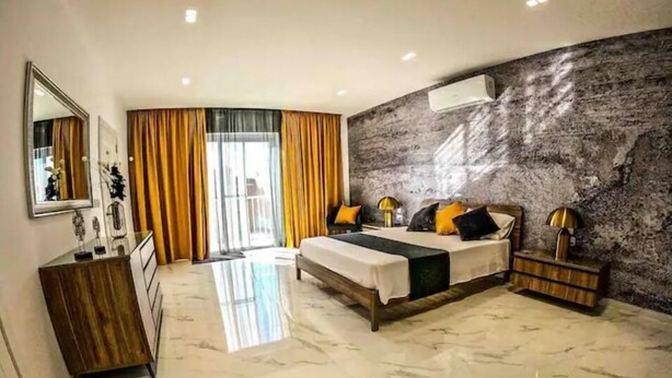 Une chambre à coucher moderne avec un sol en marbre, un lit agrémenté de plusieurs coussins, une table de chevet avec une lampe et un grand miroir.