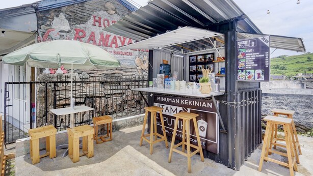 Un stand de nourriture avec un panneau affichant le menu, des tabourets en bois et un mur de pierres en arrière-plan.