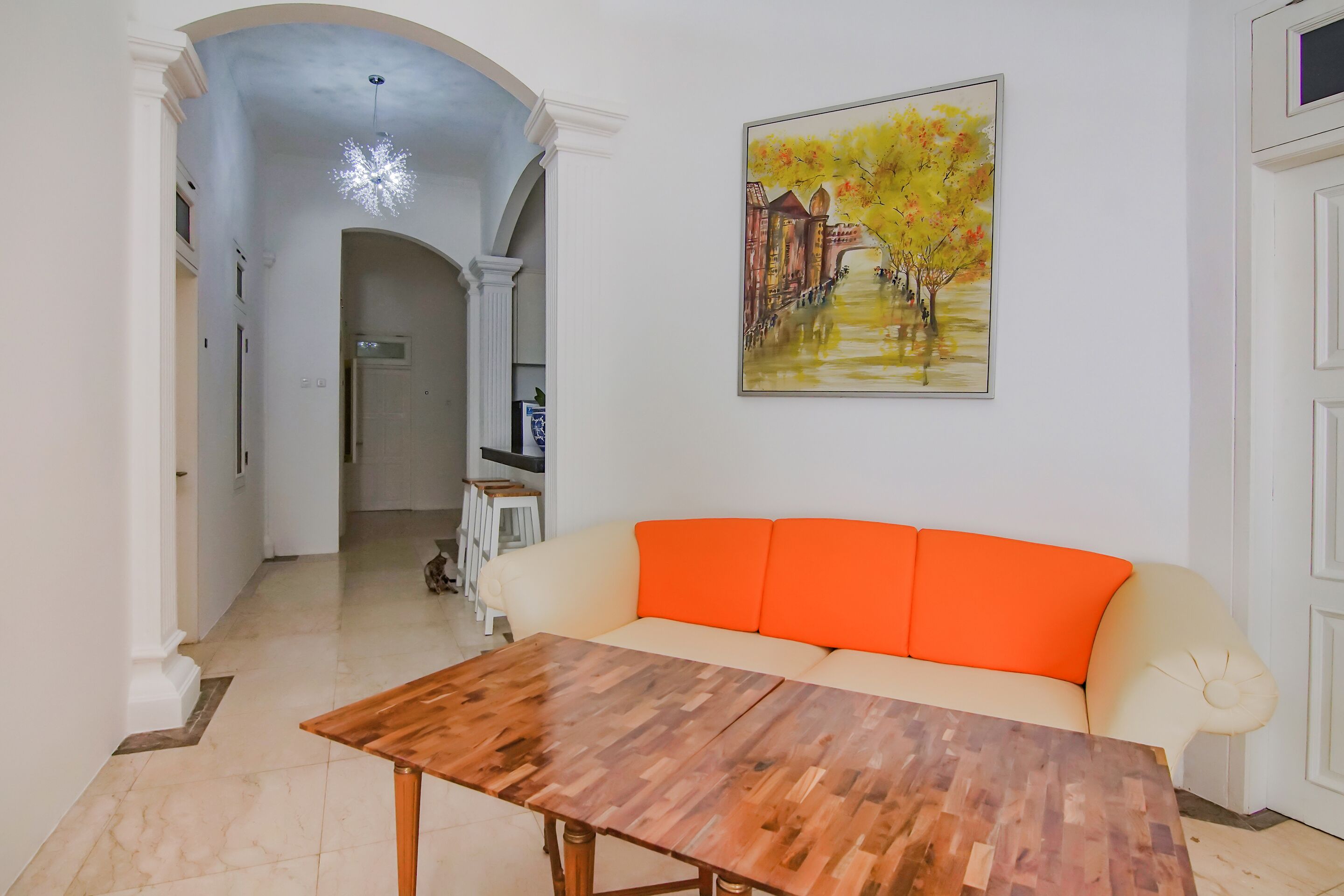 A living room with a wooden coffee table, a cream-colored sofa with orange cushions, and a painting of a tree-lined pathway on the wall.