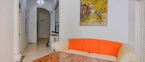 A living room with a wooden coffee table, a cream-colored sofa with orange cushions, and a painting of a tree-lined pathway on the wall.