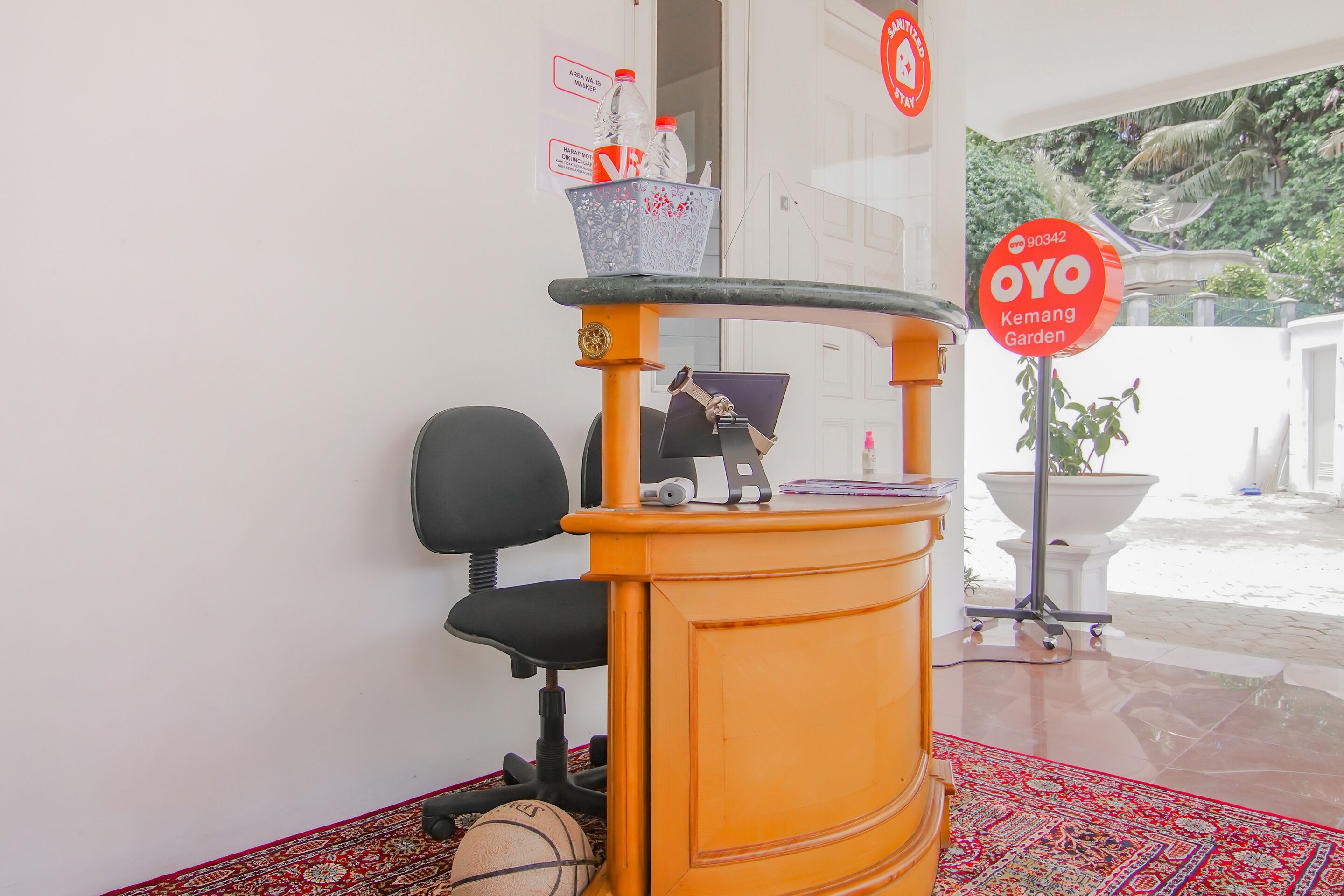 A room with a desk, chair, and a basketball on the floor. There is a sign indicating a mask requirement and a sign for OYO Kemang Garden.