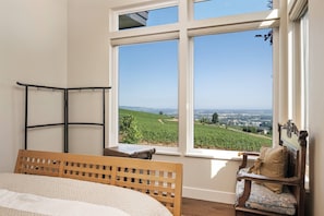 A bedroom with a large window offering a view of a vineyard and distant landscape.