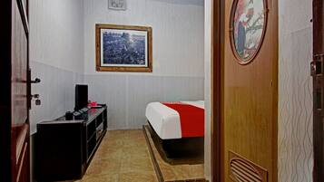 Habitación de hotel pequeña con cama, escritorio, silla, mesita, un cuadro en la pared y una puerta con panel decorativo.