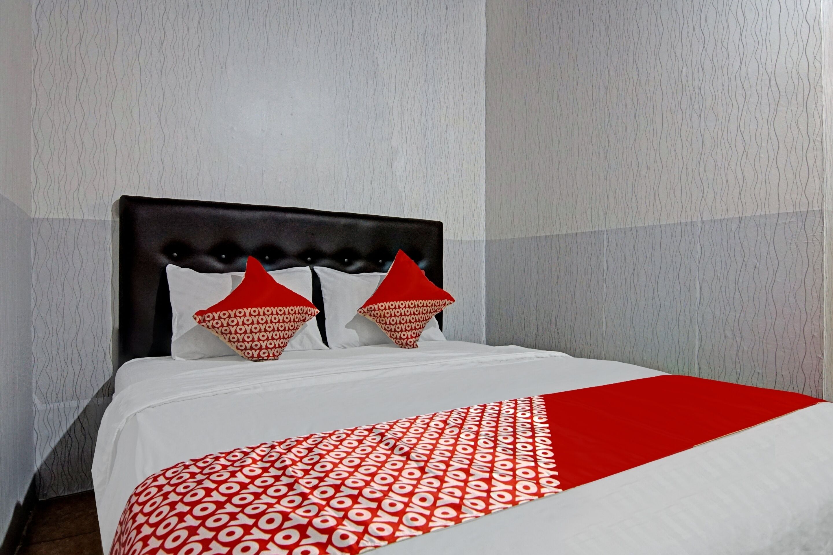 A bed with a white and red patterned bedspread, a dark headboard, and two red pillows with a geometric pattern.