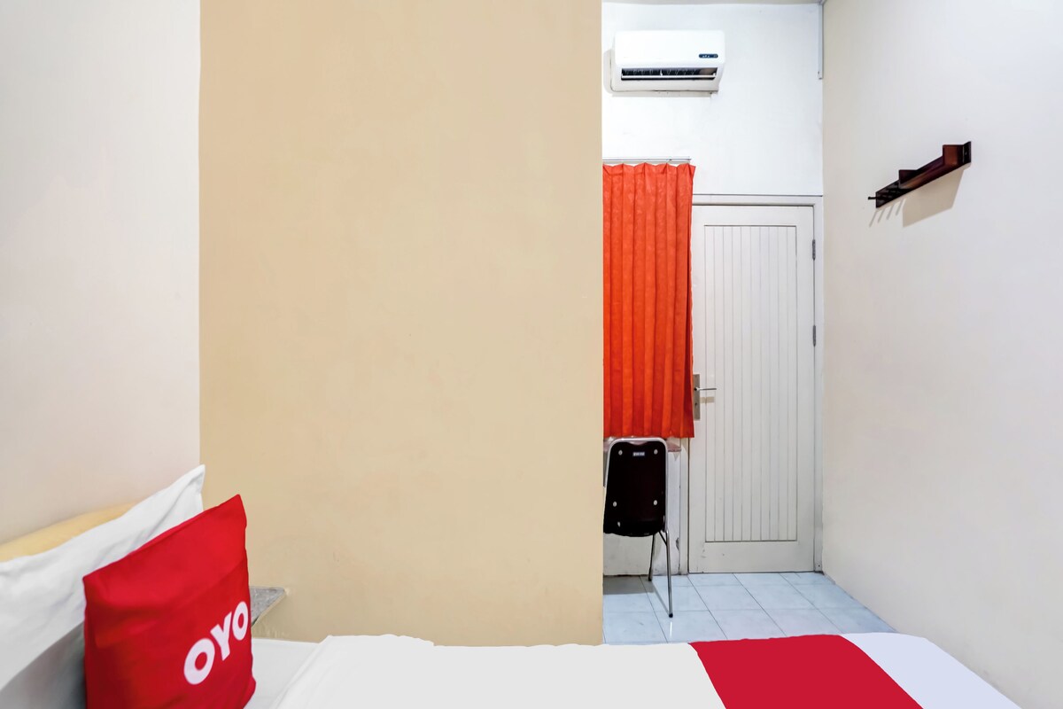 Une chambre d’hôtel avec un lit, un oreiller rouge, un oreiller blanc, un appareil de climatisation, une porte avec un rideau orange et une petite table avec une chaise.