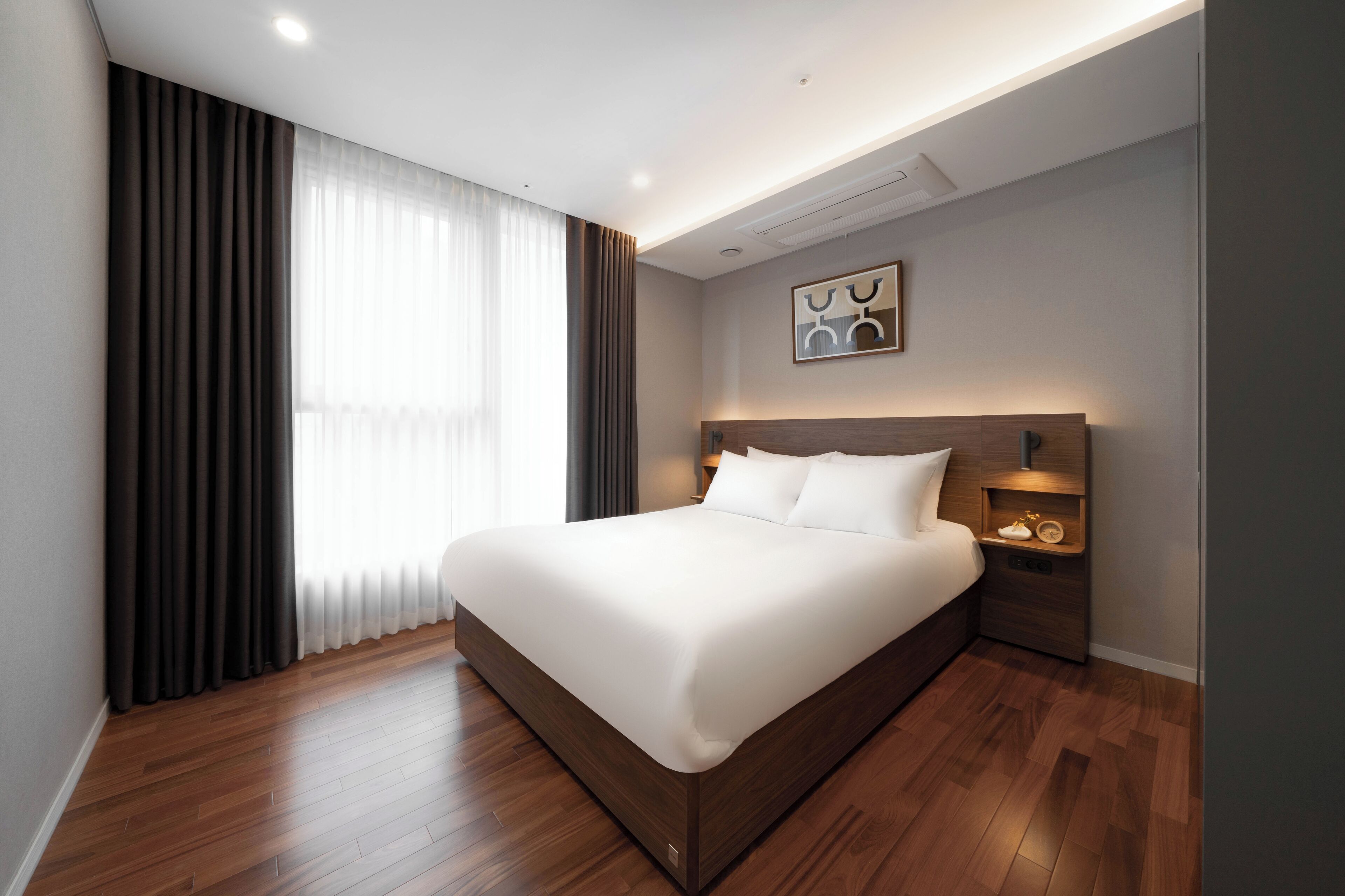 Una habitación de hotel moderna con una cama grande, suelo de madera, una mesita de noche con lámpara y vistas al exterior a través de cortinas translúcidas.