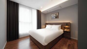 Una habitación de hotel moderna con una cama grande, suelo de madera, una mesita de noche con lámpara y vistas al exterior a través de cortinas translúcidas.