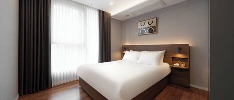 A modern hotel room with a large bed, wooden flooring, a nightstand with a lamp, and a view of the outdoors through sheer curtains.