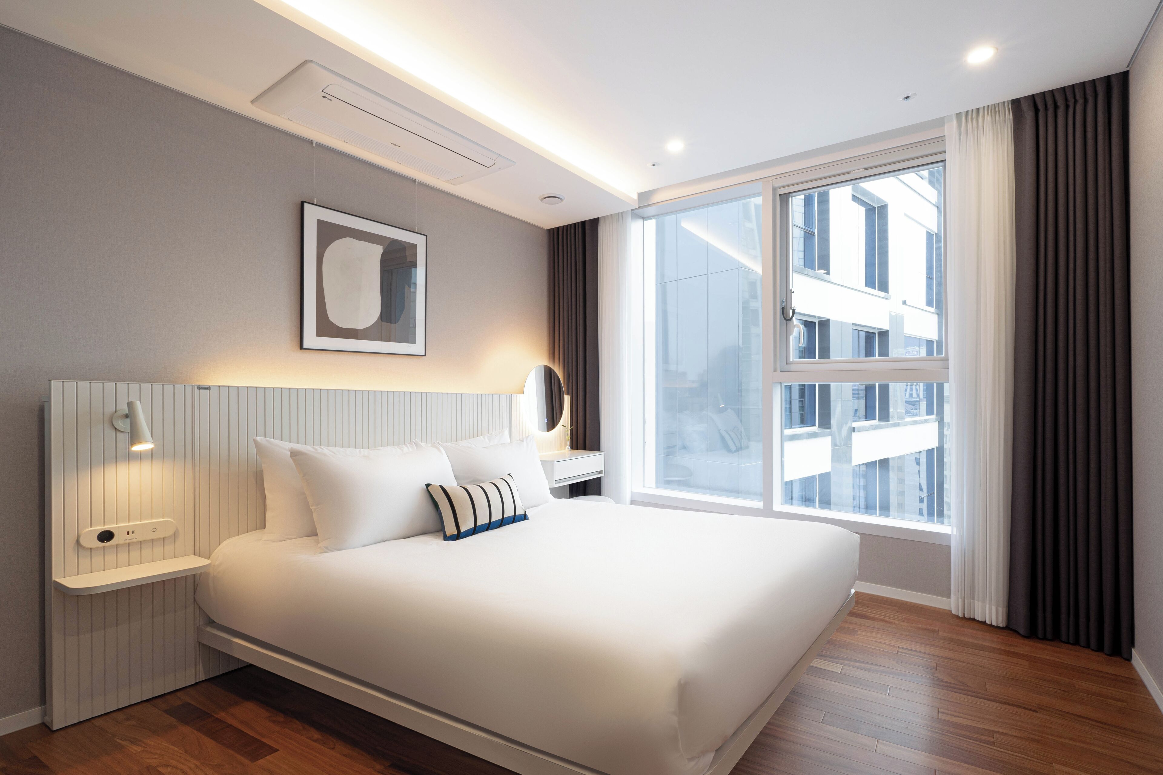 A modern hotel room with a large bed, bedside tables, a wall-mounted mirror, and a large window with curtains.
