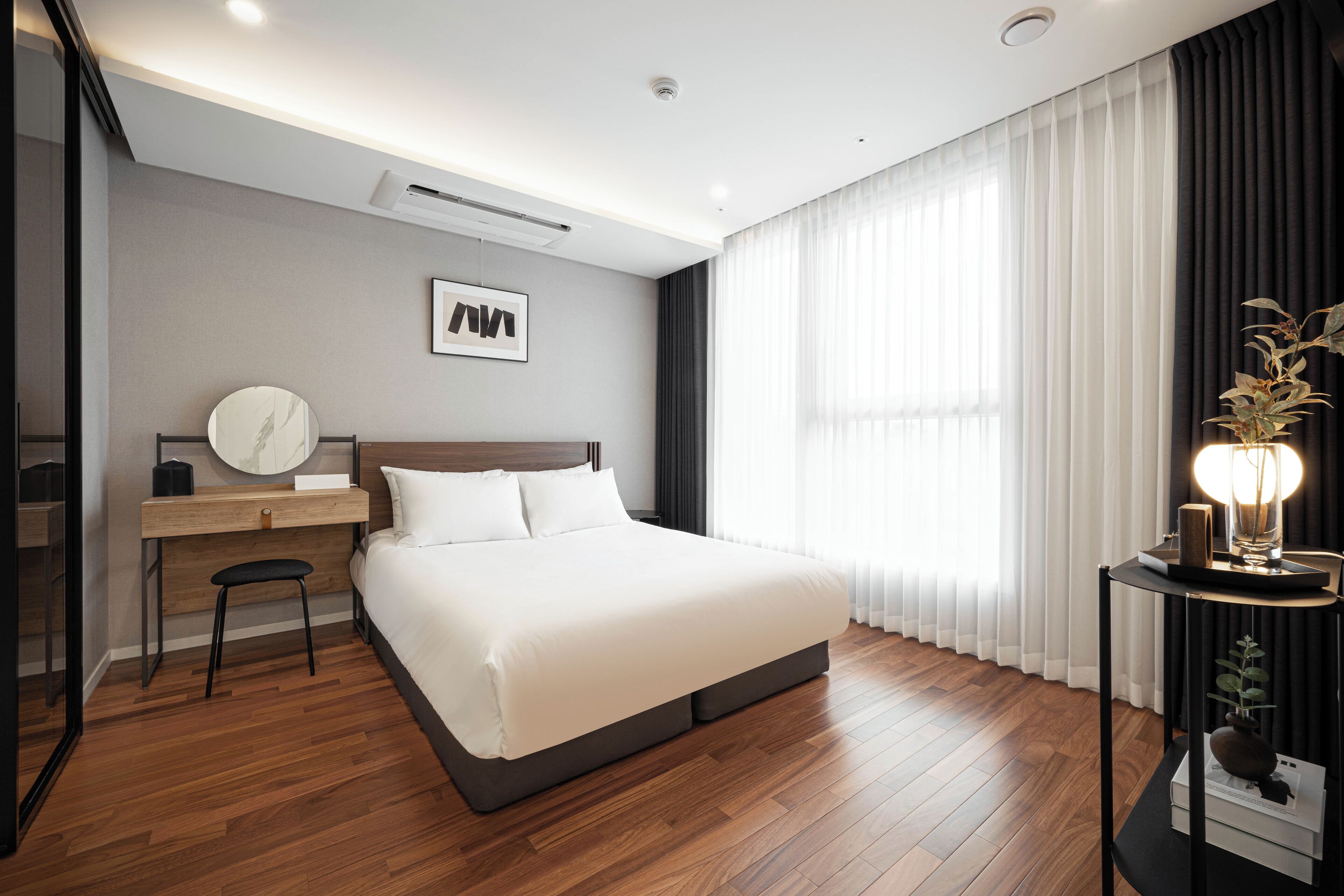 Una habitación de hotel moderna con una cama grande, un escritorio con silla, un espejo redondo y una mesita de noche con lámpara.