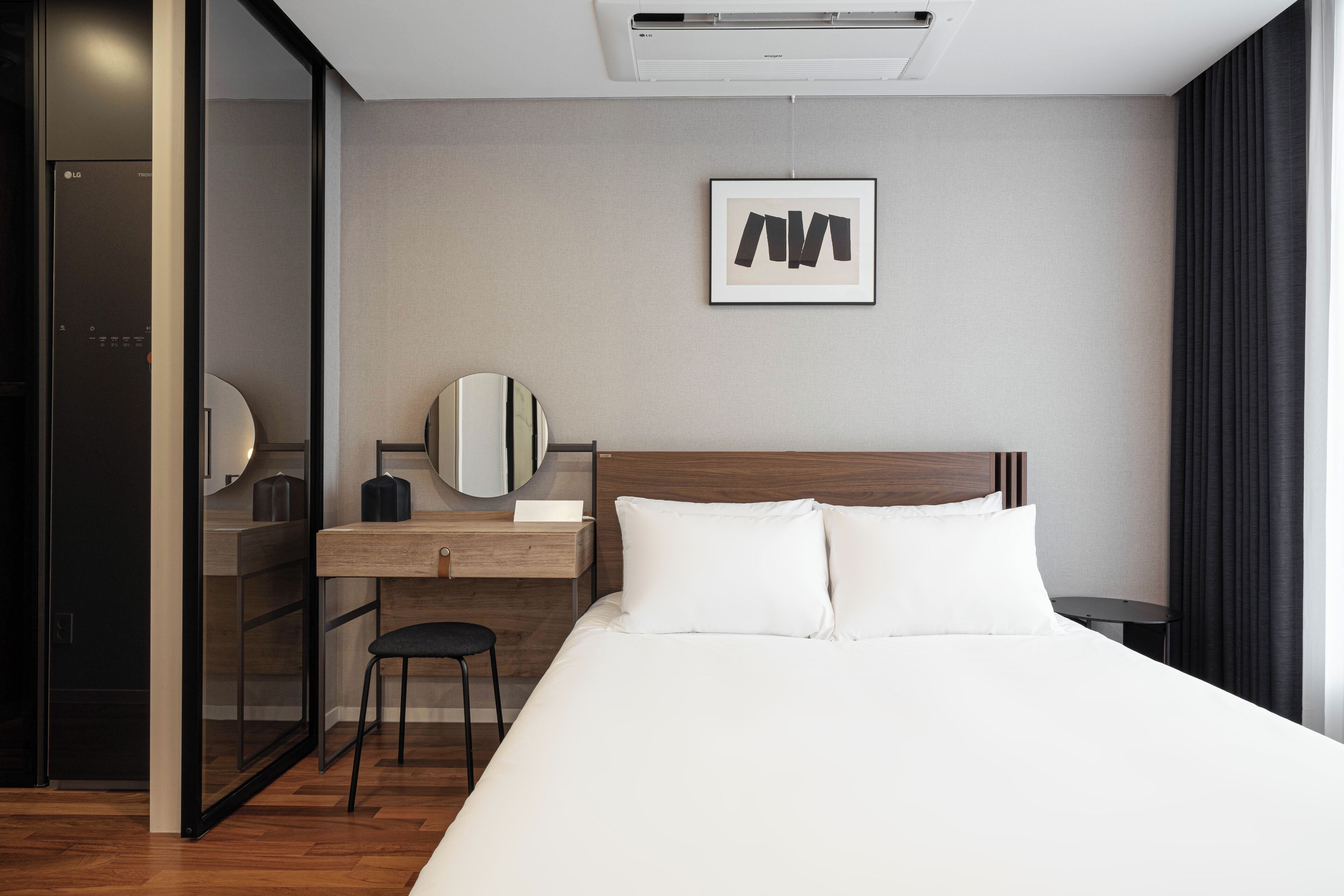 A modern hotel room with a bed, a desk with a chair, a mirror, and a framed artwork on the wall.