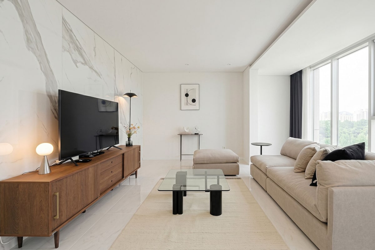 A modern living room with a flat-screen TV, wooden cabinet, glass coffee table, beige sofa, and large windows.