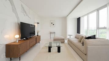A modern living room with a flat-screen TV, wooden cabinet, glass coffee table, beige sofa, and large windows.