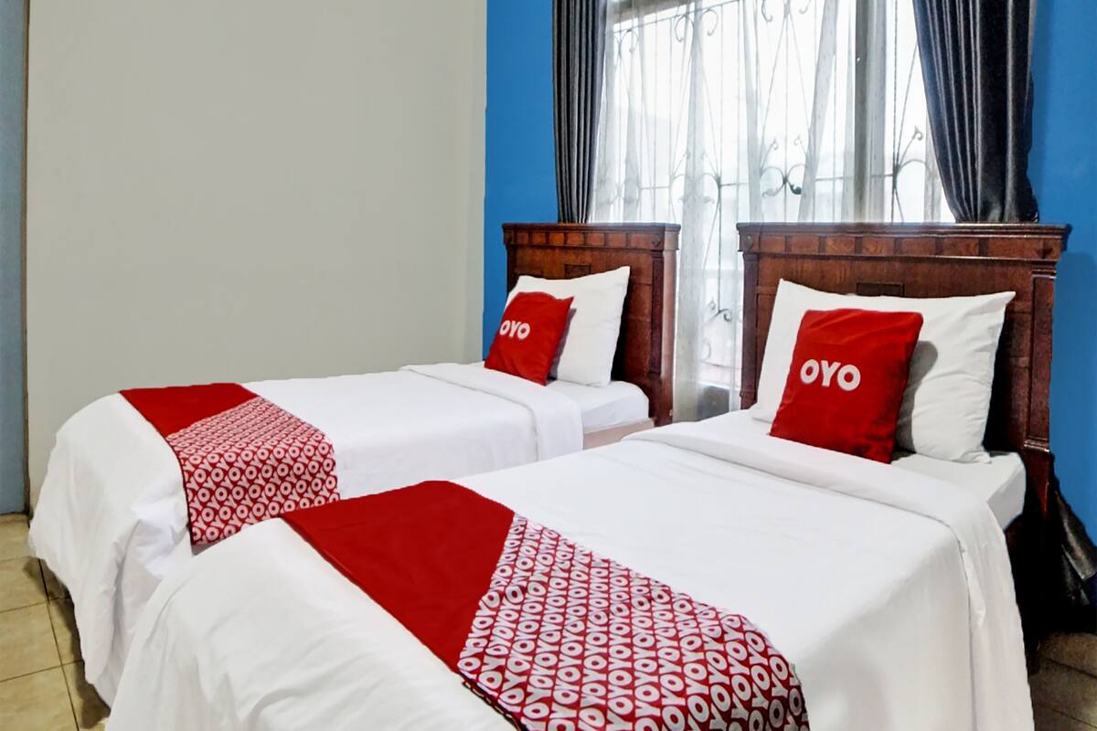 Hotellihuoneessa on kaksi sänkyä, joissa on molemmissa punavalkoinen kuviollinen peitto ja samanväriset tyynyt. Huoneessa on puinen sängynpääty, ikkuna, jossa on verhot, ja sininen seinä.