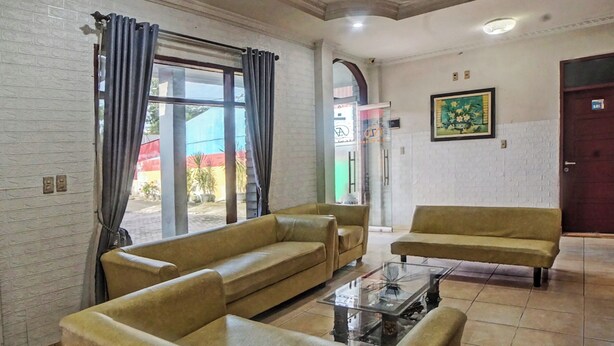 A living room with two sofas and a coffee table, a large window with curtains, a door with a sign, and a framed picture on the wall. - Hotel O Serena Anggrek (Medan)