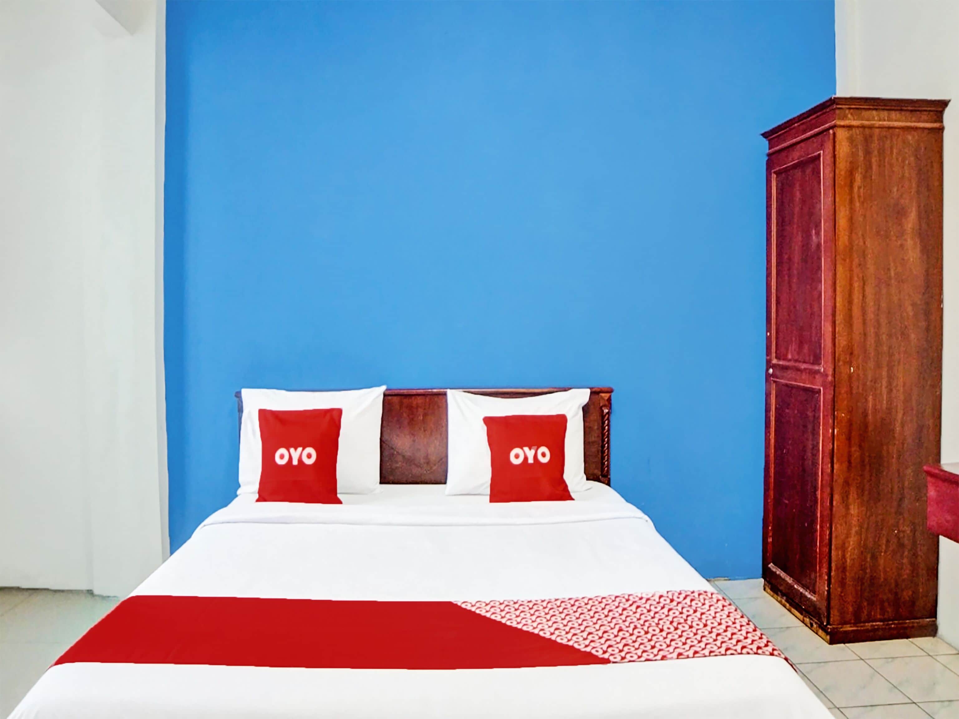 A bed with a white and red bedspread, two pillows with 'OYO' branding, and a wooden wardrobe.