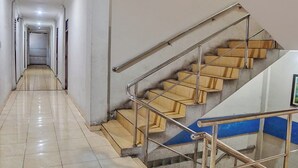 A long, narrow hallway with a staircase on the right. The hallway has a tiled floor and white walls. The staircase has wooden steps and a metal railing. - Hotel O Serena Anggrek (Medan)