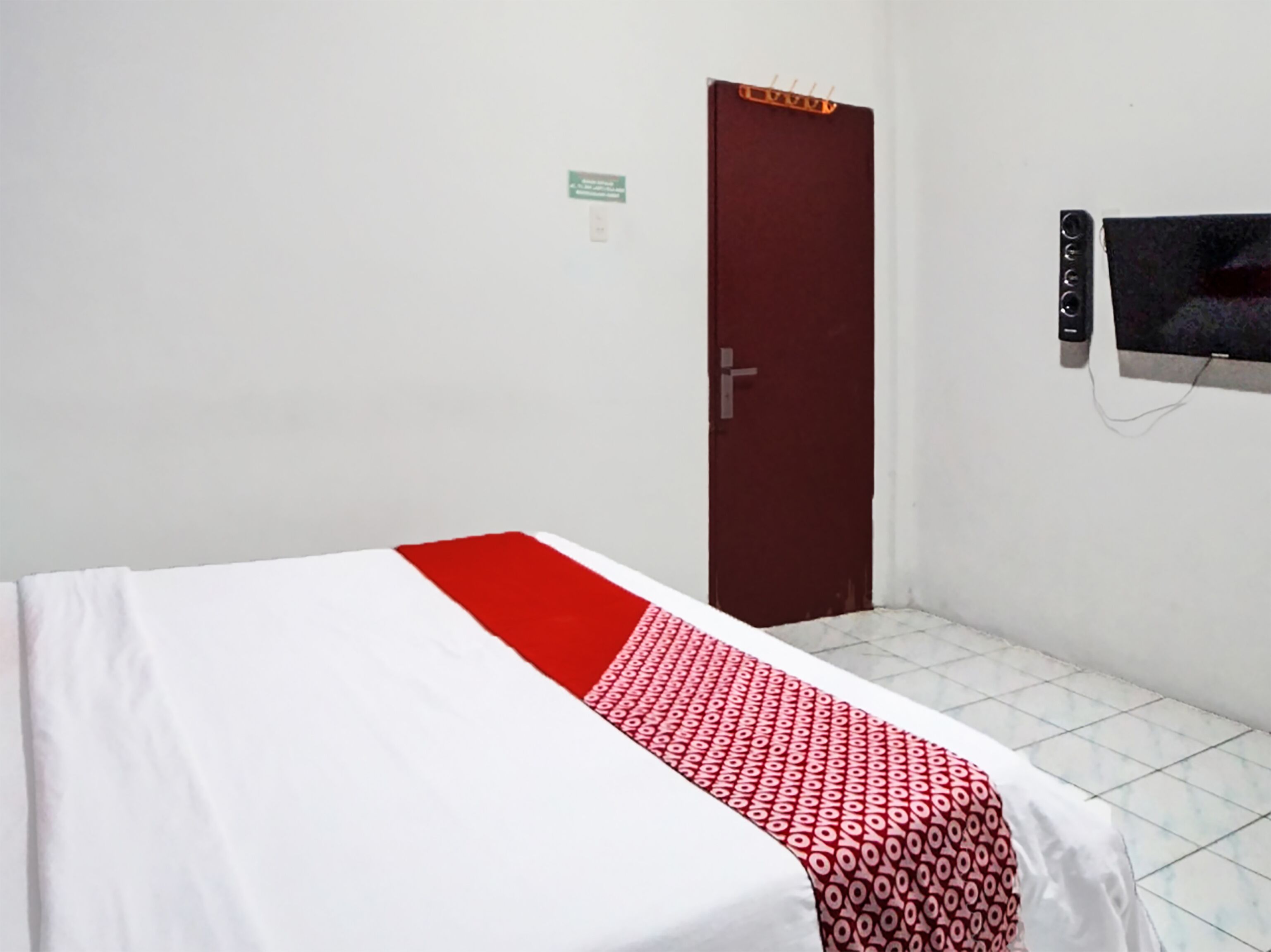 Habitación con cama individual, colcha roja y blanca, puerta cerrada y televisor montado en la pared.