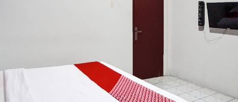 Habitación con cama individual, colcha roja y blanca, puerta cerrada y televisor montado en la pared.