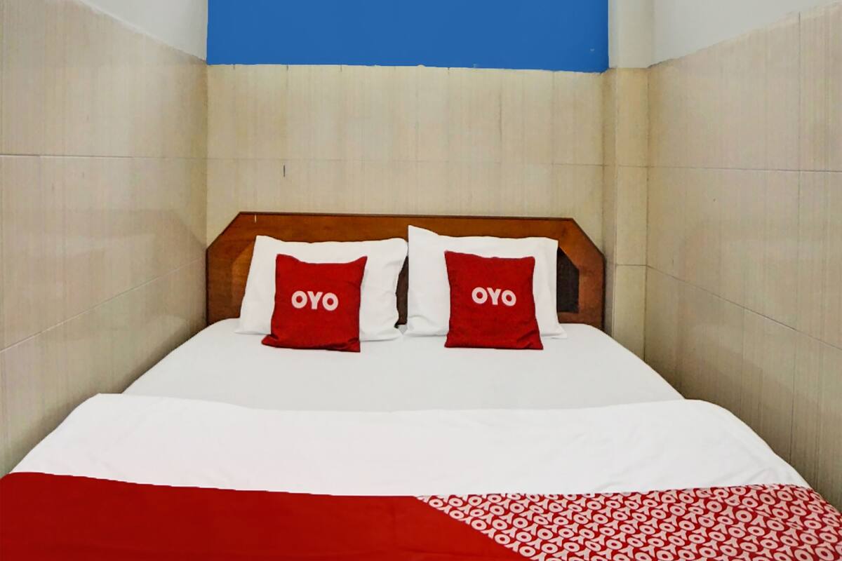 Hotellihuone, jossa on sänky, kaksi punaista tyynyä, joissa on teksti ”OYO”, valkoiset vuodevaatteet ja punavalkoinen kuviollinen peitto.