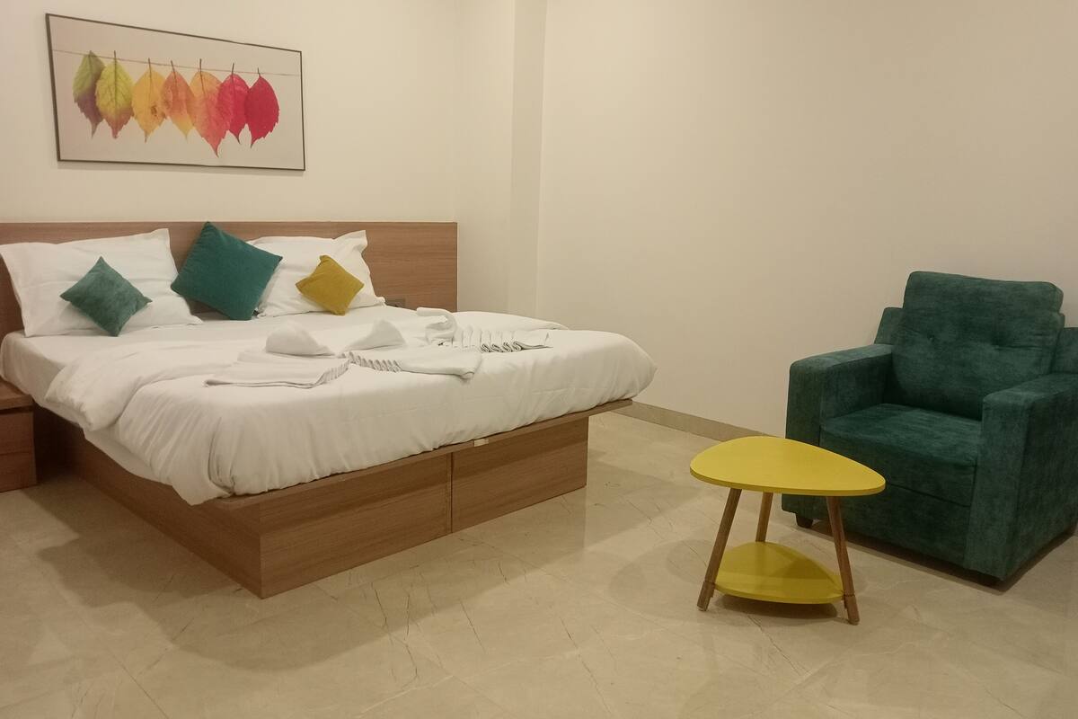 Una cama bien hecha con almohadas, una mesita de noche, una silla y una obra de arte en la pared.
