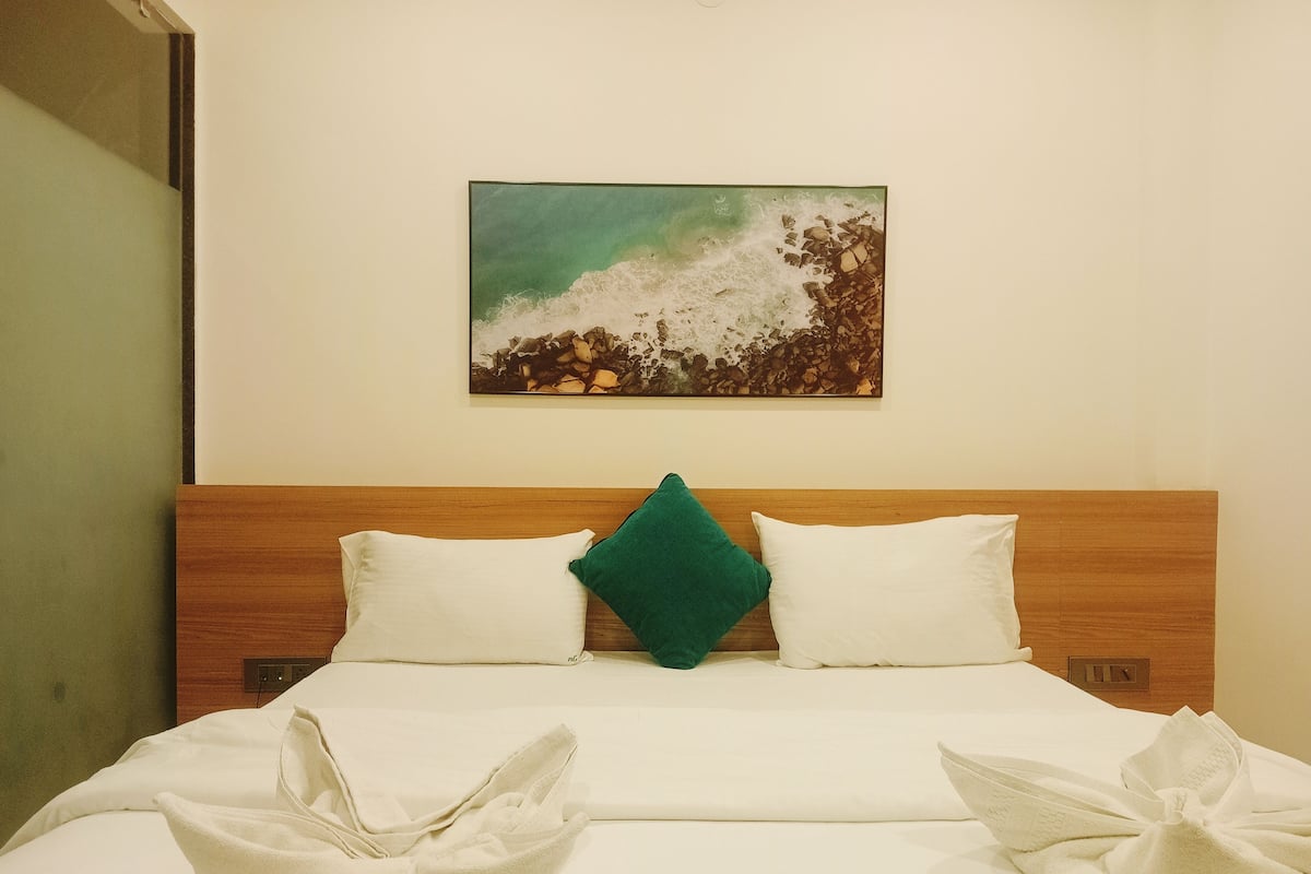 Habitación de hotel con cabecera de madera, dos almohadas blancas y una almohada decorativa verde, junto a un cuadro de una playa.