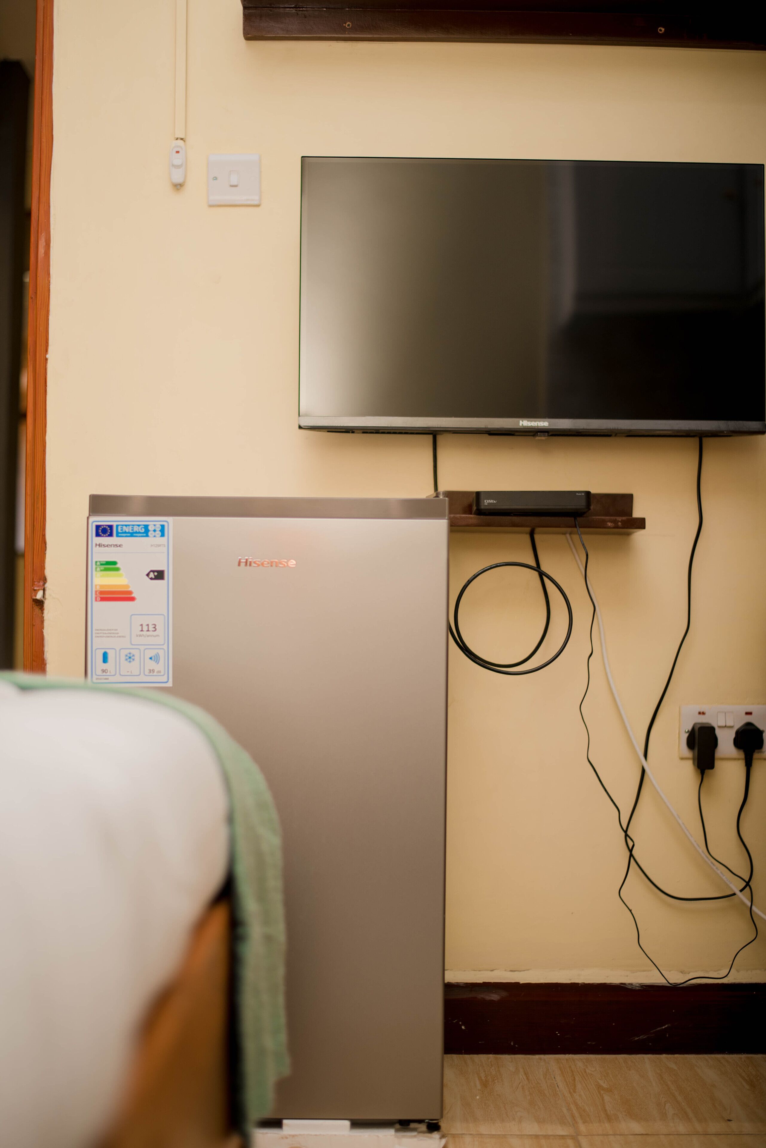 Habitació compacta amb llit, nevera Hisense, televisió penjada i preses elèctriques visibles.