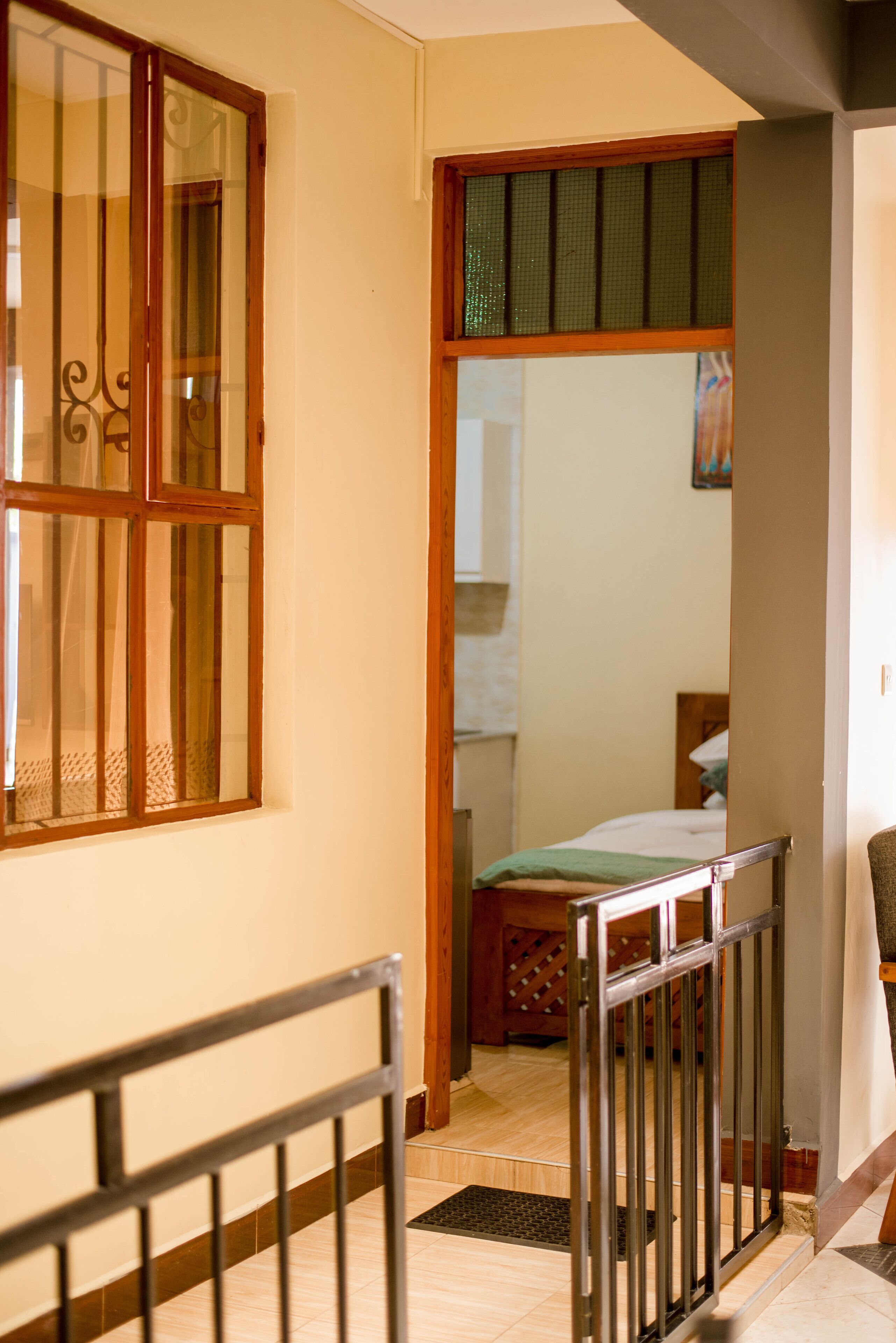 A hallway with a wooden door, a window with bars, and a bed in the background.