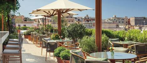 An outdoor patio with multiple tables and chairs, surrounded by potted plants and umbrellas.