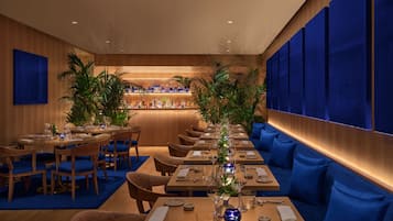 Un restaurant avec des tables en bois et des banquettes recouvertes d’un tissu bleu, un coin bar à la décoration bleue et des plantes en pot.