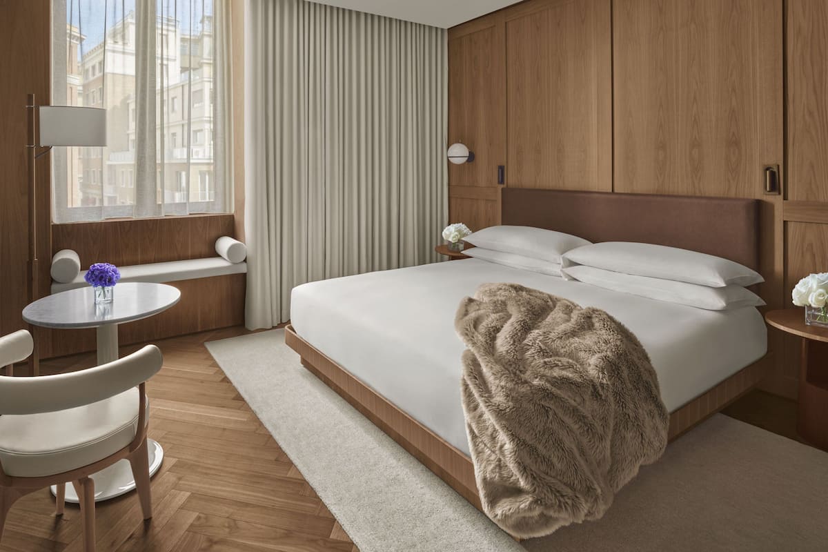 Habitació d'hotel amb un llit gran, una taula de nit, una cadira, una finestra amb cortines i vista a edificis a l'exterior.