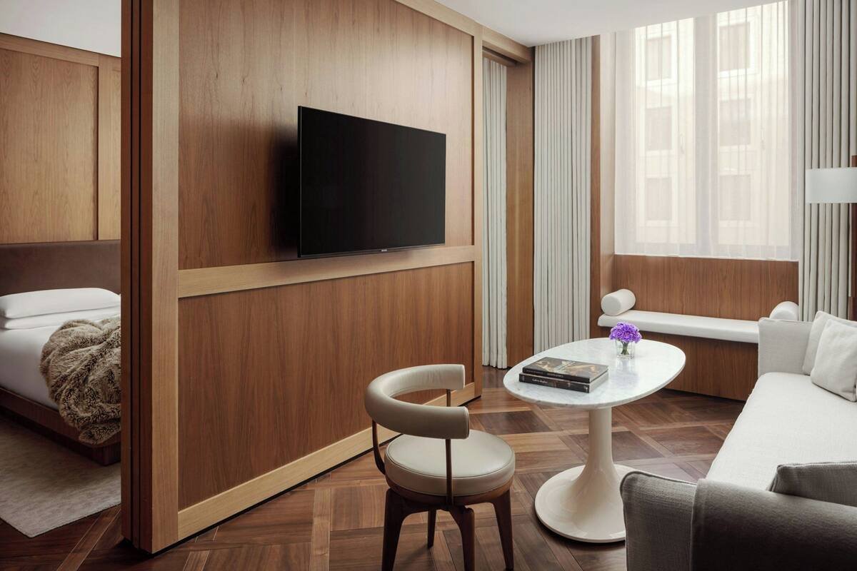 Habitació d'hotel moderna amb un televisor de pantalla plana fixat a un panell de fusta, un sofà blanc, una taula de centre rodona amb un llibre i un vas, i un paviment de fusta.