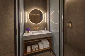 A modern bathroom with a marble sink, a round mirror with integrated lighting, a vanity with a shelf for towels, and a glass shower enclosure.