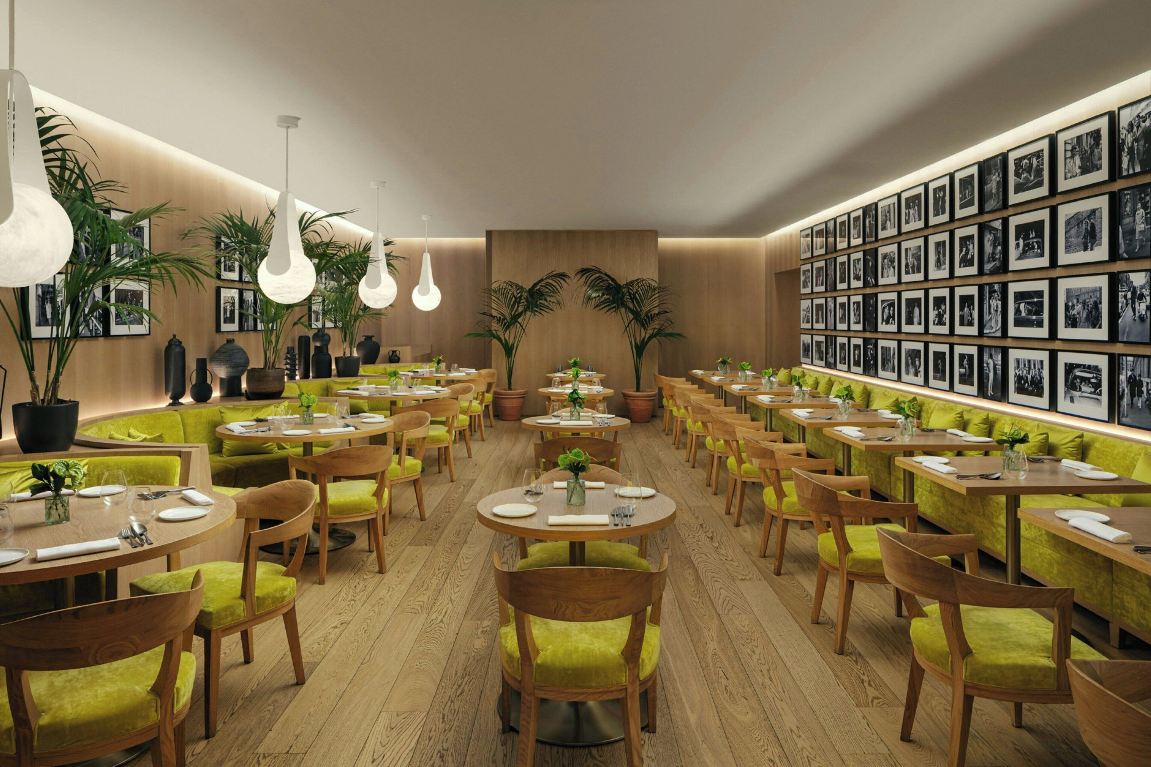 A restaurant with round tables, green cushioned chairs, potted plants, and a wall of framed photographs.