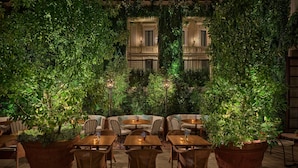 An outdoor seating area with tables and chairs, surrounded by large potted plants and a building with balconies in the background. - The Rome EDITION (Rome)