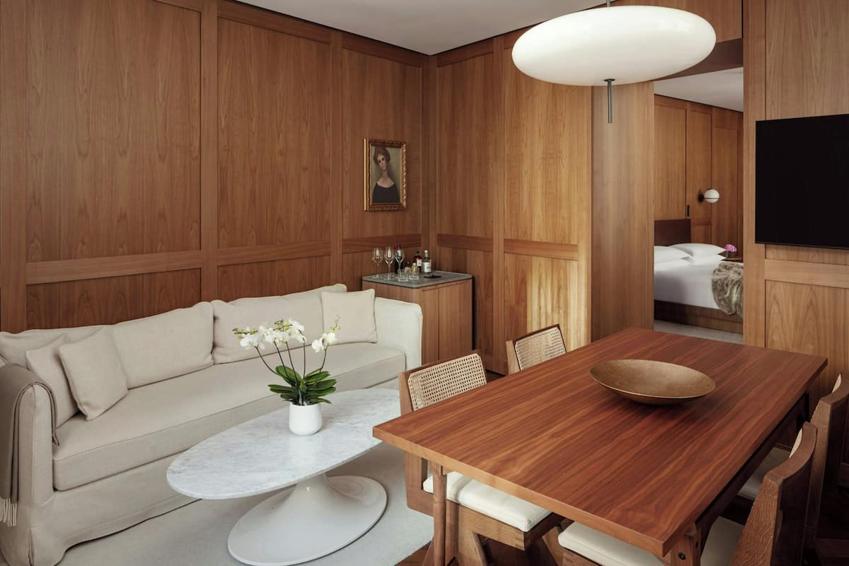 Habitació d'hotel moderna amb paret de panells de fusta, un sofà, una taula de dinar amb cadirees, una petita taula auxiliar amb un vas i gots de vi, i un televisor fixat a la paret.
