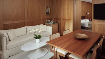A modern hotel room with a wooden panel wall, a sofa, a dining table with chairs, a small side table with a vase and wine glasses, and a television mounted on the wall.