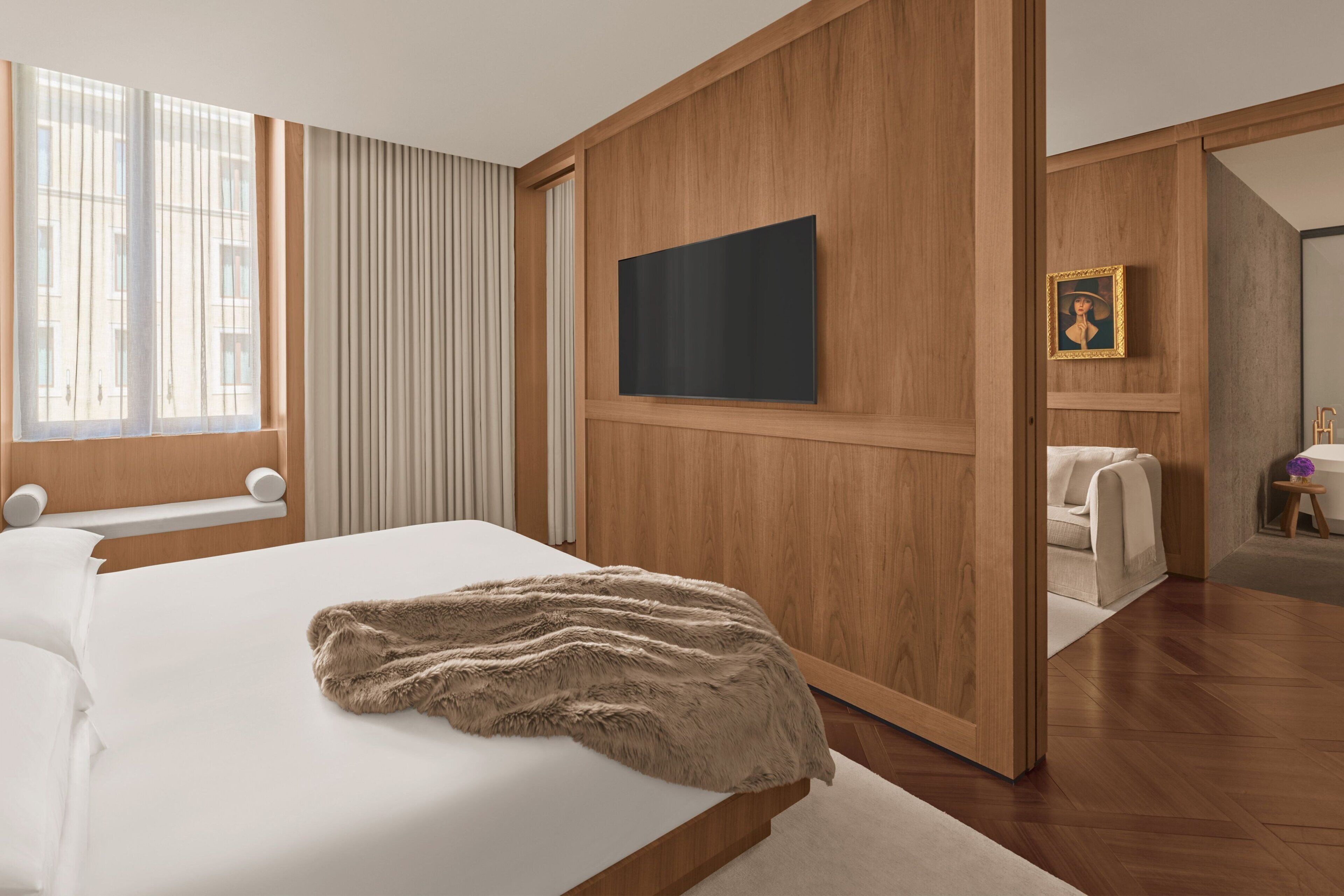 Una habitación de hotel moderna con una cama grande, paneles de madera, un televisor de pantalla plana, un cuadro enmarcado y vistas de la ciudad a través de amplias ventanas.