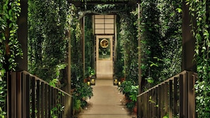 A corridor with a wooden floor, flanked by vertical metal railings and potted plants, leading to a door with a circular emblem. - The Rome EDITION (Rome)