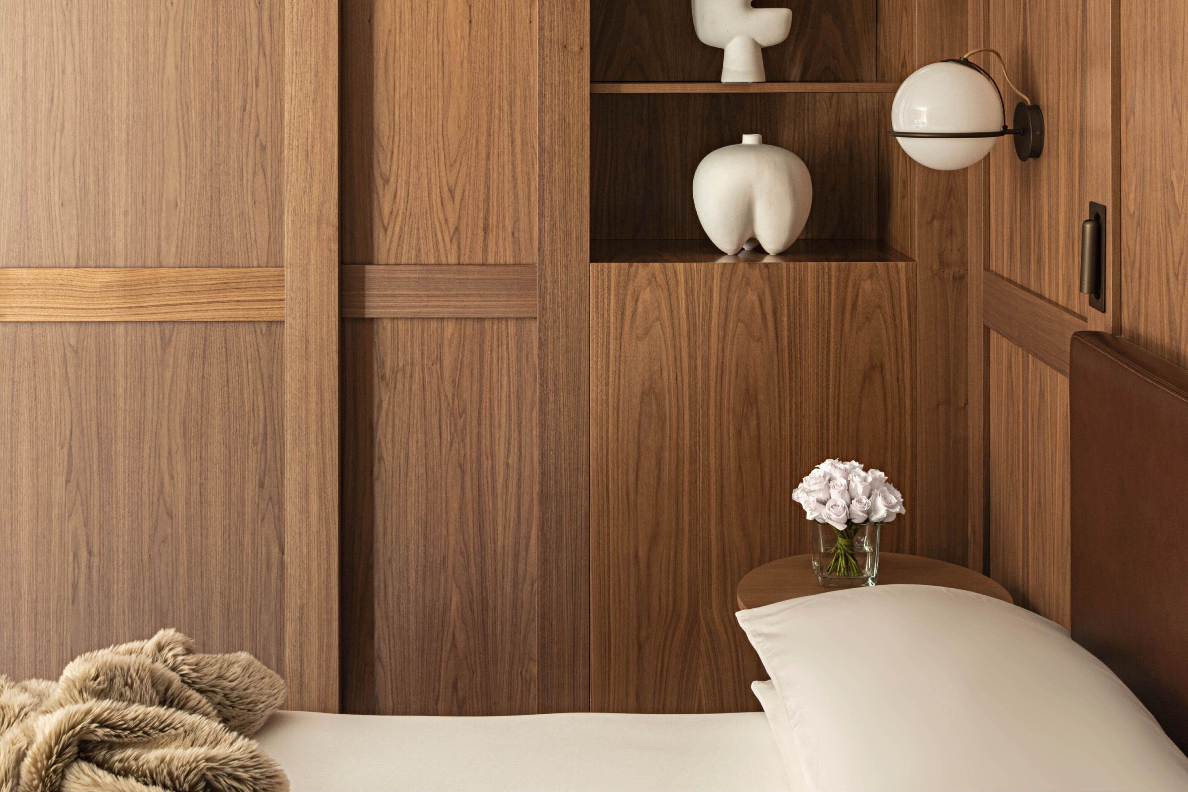 Une pièce en bois avec un lit, une table de chevet ornée d’un vase de fleurs et une lampe fixée au mur.