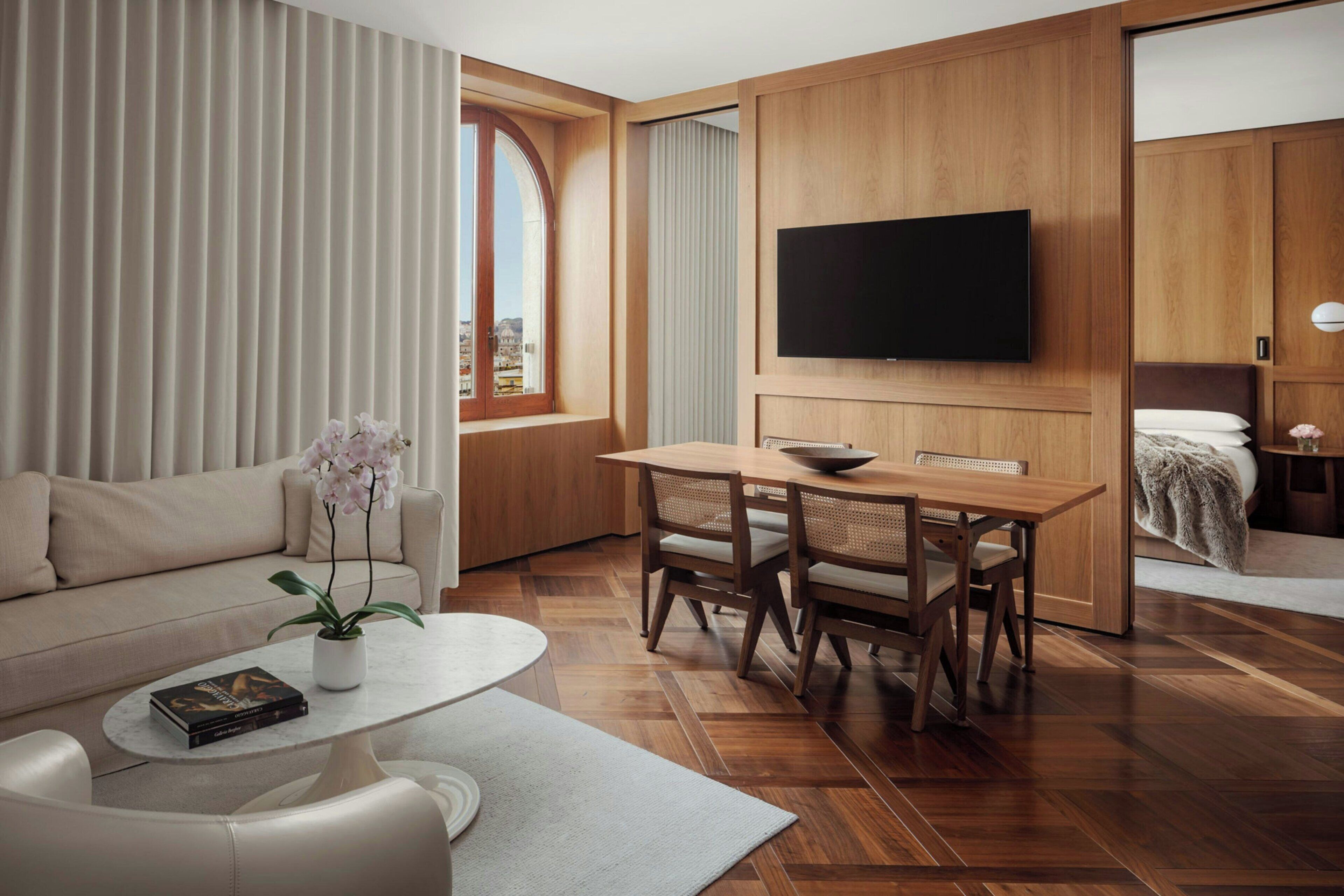 A modern hotel room with a wooden floor, a flat-screen TV mounted on the wall, a dining area with a table and chairs, a sofa, and a window with curtains.