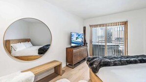 A modern bedroom with a round mirror, a bed, a TV, a wooden bench, and a balcony with a view of the city. - Luxe DV Condo at Snow Park (Park City)