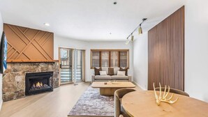 A modern living room with a stone fireplace, a sofa, a coffee table, and a dining table with a decorative antler. - Luxe DV Condo at Snow Park (Park City)