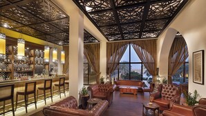 A spacious lounge area with leather sofas and armchairs, a bar with a variety of bottles, and large windows offering a view of the outdoors. - Aldhafra Resort, Vignette Collection by IHG (Madinat Zayed)