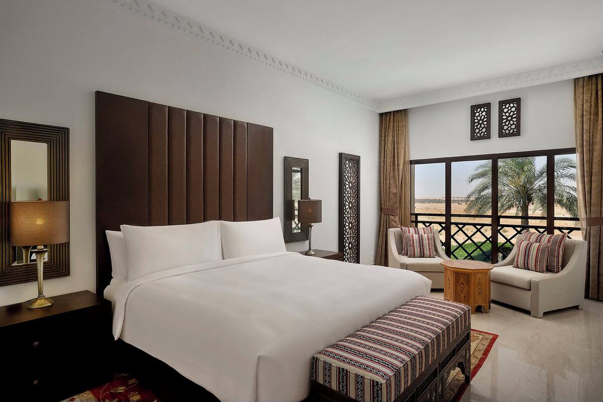 A hotel room with a large bed, a sofa, a small table, and a view of palm trees through the window.