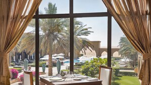 A dining area with a table set for two, overlooking a poolside with palm trees and lounge chairs. - Aldhafra Resort, Vignette Collection by IHG (Madinat Zayed)