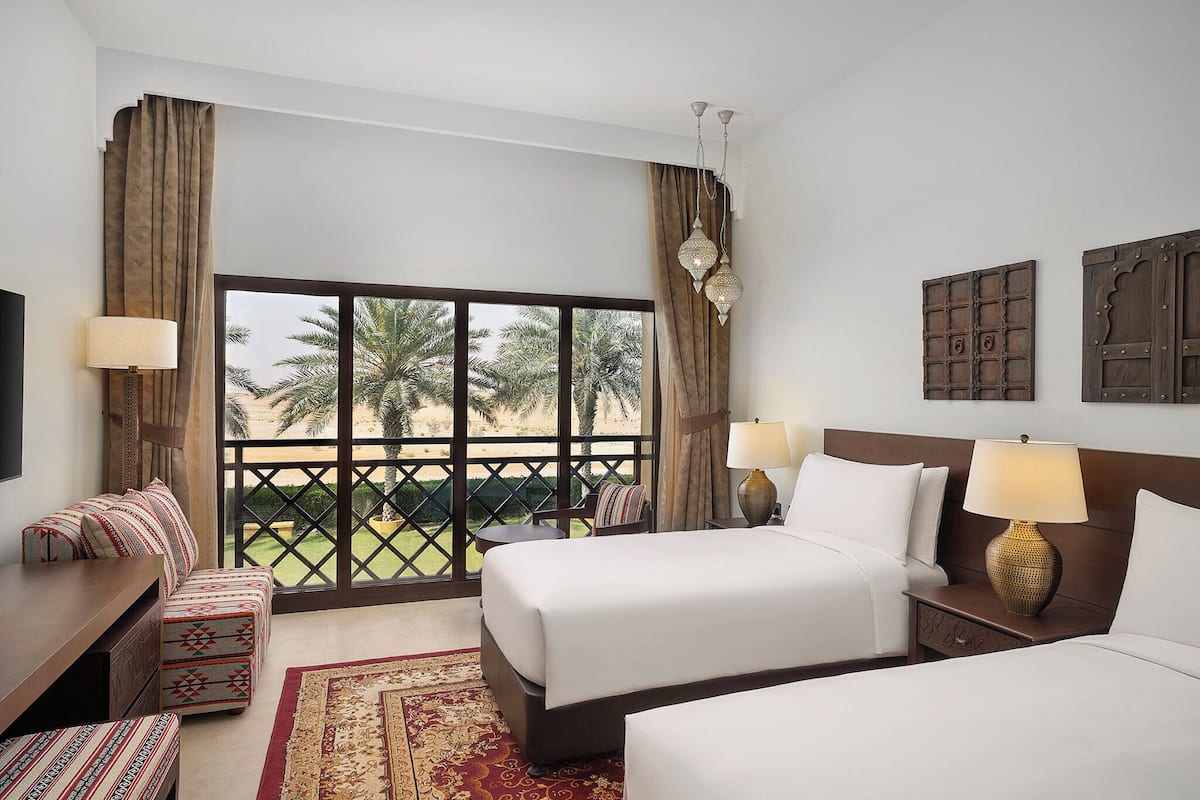 A hotel room with two beds, a sofa, a small table, and a view of palm trees through large windows.